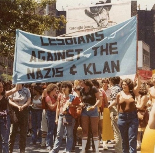 The photograph depicts a vibrant outdoor scene, likely a protest or demonstration, filled with a large crowd of people. The central focus is a light blue banner held aloft by several individuals.  The banner boldly proclaims, "LESBIANS AGAINST THE N***S & K**N" in large, dark lettering. The women holding the banner and surrounding participants have a mix of hairstyles and clothing styles typical of the 1970s. Their attire ranges from jeans and t-shirts to more fitted tops and shorts. Many wear knee-high socks. One woman in the center is wearing a black short-sleeved shirt and denim shorts. Others wear loose-fitting tops, jeans and skirts. The overall style reflects a casual, yet determined, attitude. Several participants appear to be wearing brightly colored shirts or shirts with patterns. The crowd is densely packed, and the faces of many individuals are clearly visible, expressing a mix of seriousness and determination. The atmosphere feels energized and politically charged, reflecting the seriousness of the protest. The mood is one of defiance and solidarity. In the background, a large billboard is partially visible. It features a black and white image of a man wearing a cowboy hat, with text that seems to advertise something. The urban setting suggests a city street scene, with buildings in the background. The area appears well-lit, indicating that the photograph was taken during daylight hours. Trees are also visible in the upper-left part of the image, suggesting it is a warm-weather day. Overall, the image is a powerful snapshot of a political event, capturing the energy and message of a lesbian protest against hate groups. This photograph's message centers strongly on the power of protest, solidarity and counter-protest to hateful groups and ideologies.